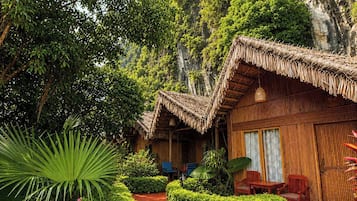 A traditional bamboo hut with a thatched roof, surrounded by lush greenery and a red-tiled pathway.