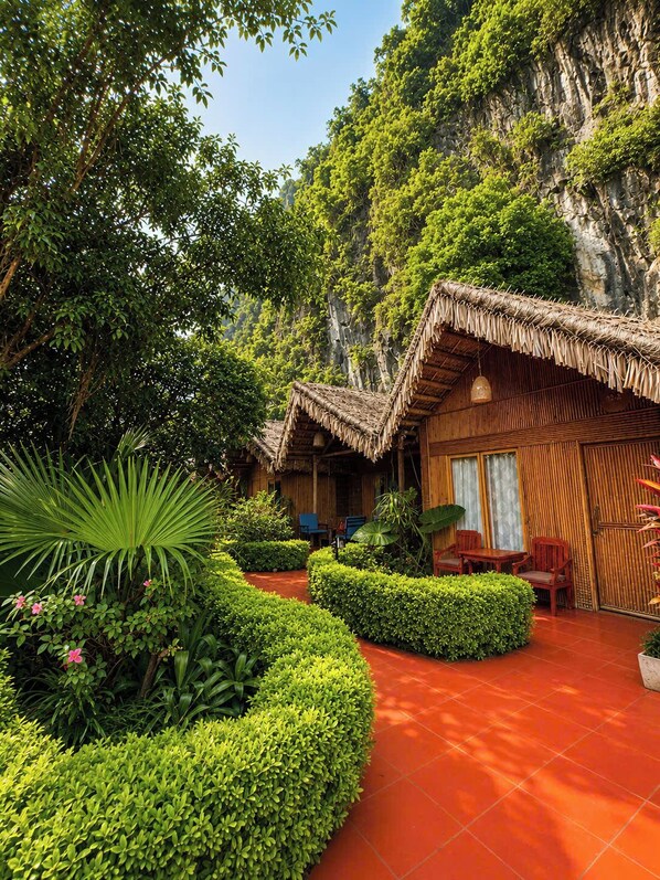 A traditional bamboo hut with a thatched roof, surrounded by lush greenery and a red-tiled pathway.
