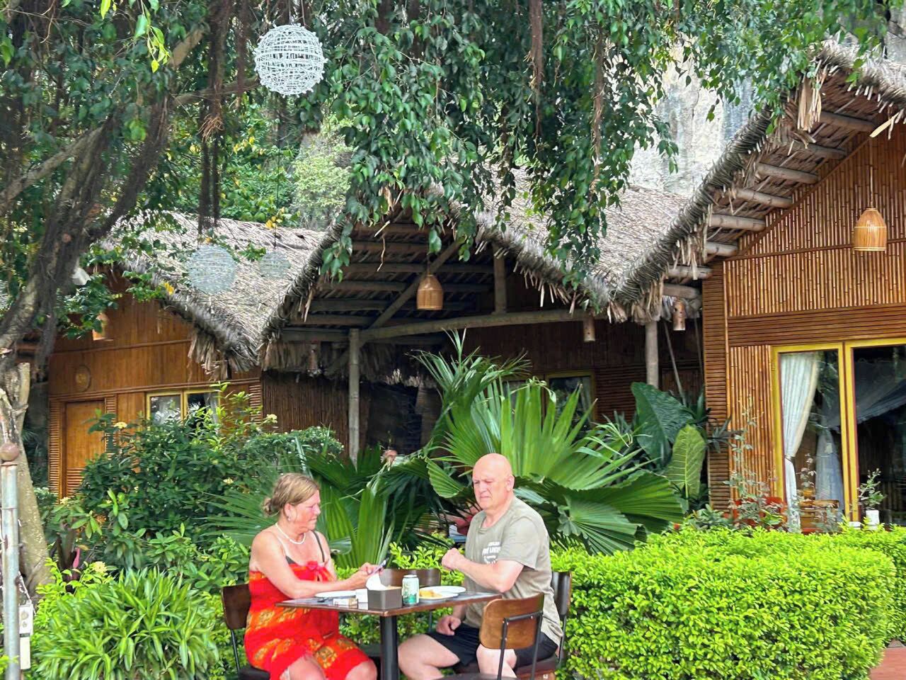 A couple dining at an outdoor table with a thatched roof and bamboo walls, surrounded by lush greenery.