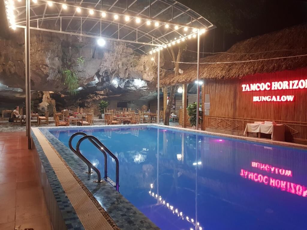 A pool area with a thatched roof structure, wooden furniture, and a neon sign reading 'TAMCOC HORIZON BUNGALOW'.