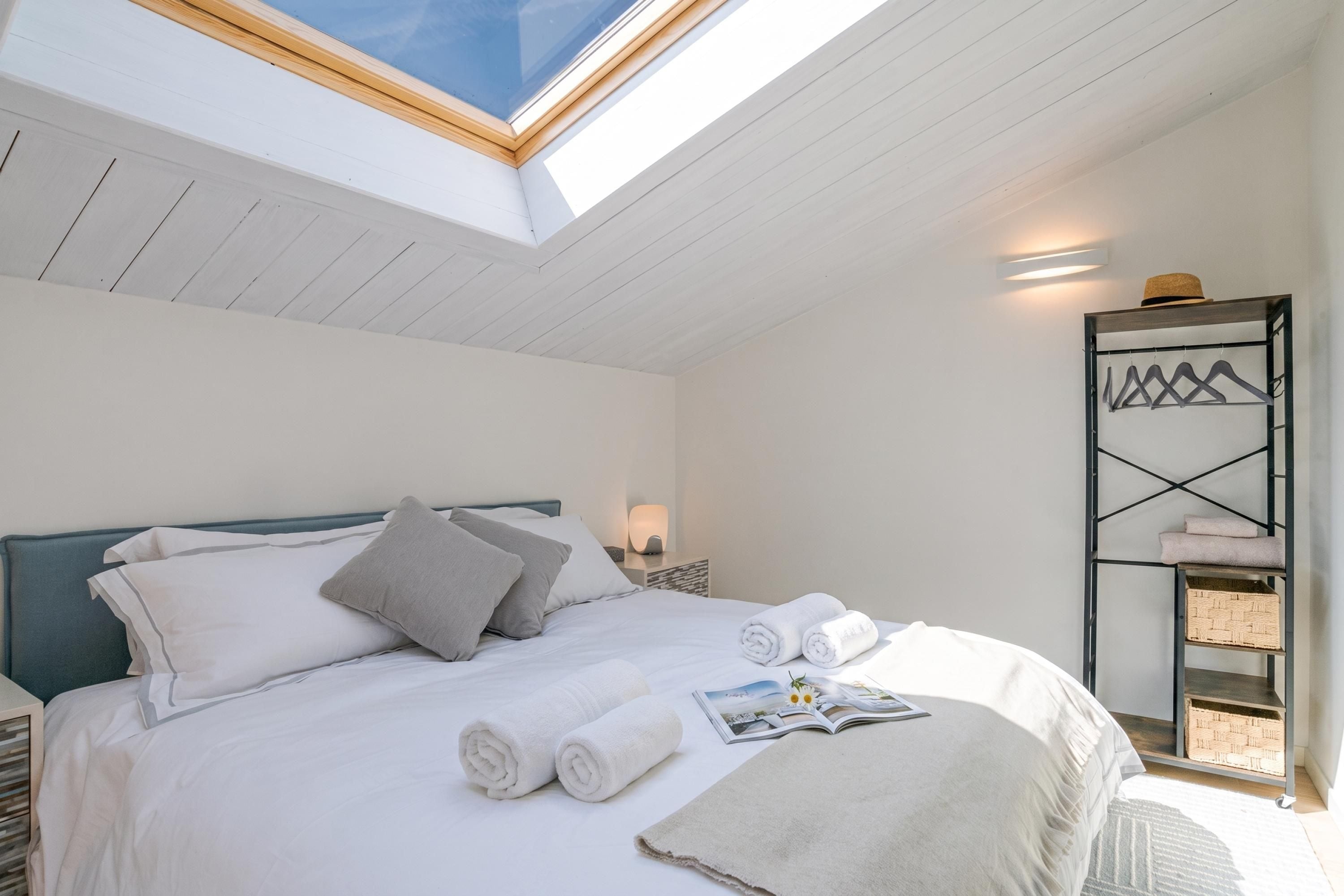 Una camera da letto con un lucernario, un letto con cuscini, asciugamani arrotolati, una rivista e uno scaffale a parete con cesti.