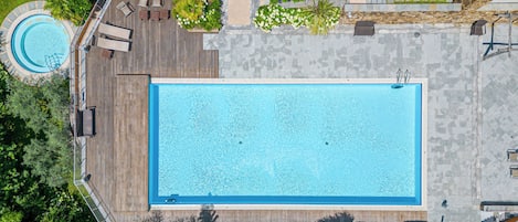 Vista aérea de uma piscina retangular com um deck de madeira ao redor, uma piscina circular menor e diversas áreas verdes.