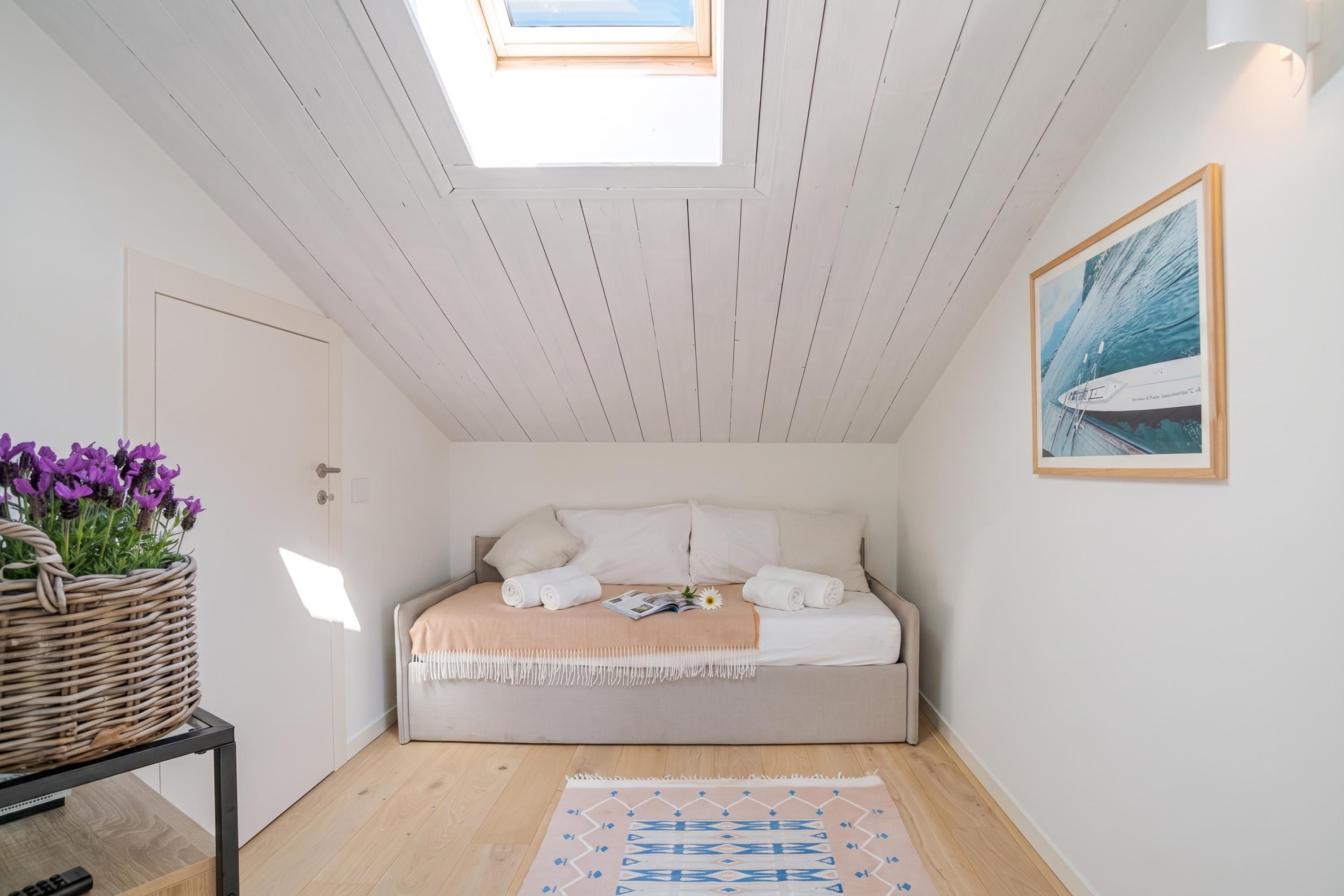 Una camera da letto con un lucernario, un letto con lenzuola bianche, un quadro appeso al muro e un cesto intrecciato con fiori viola.