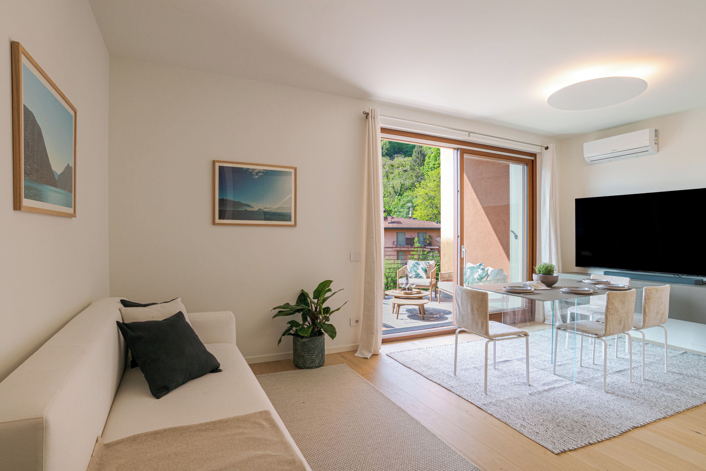 A modern living room with a sofa, a dining table with chairs, a TV, and a large window with curtains.