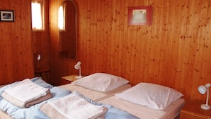 Une pièce aux murs lambrissés, avec trois lits, chacun agrémenté d’un oreiller blanc et d’une serviette pliée. On y trouve deux tables de chevet avec des lampes, une fenêtre et un tableau encadré au mur.