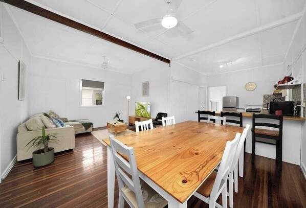 A spacious living area with a wooden dining table, white chairs, a sofa, and a kitchen area with a stove and cabinets.