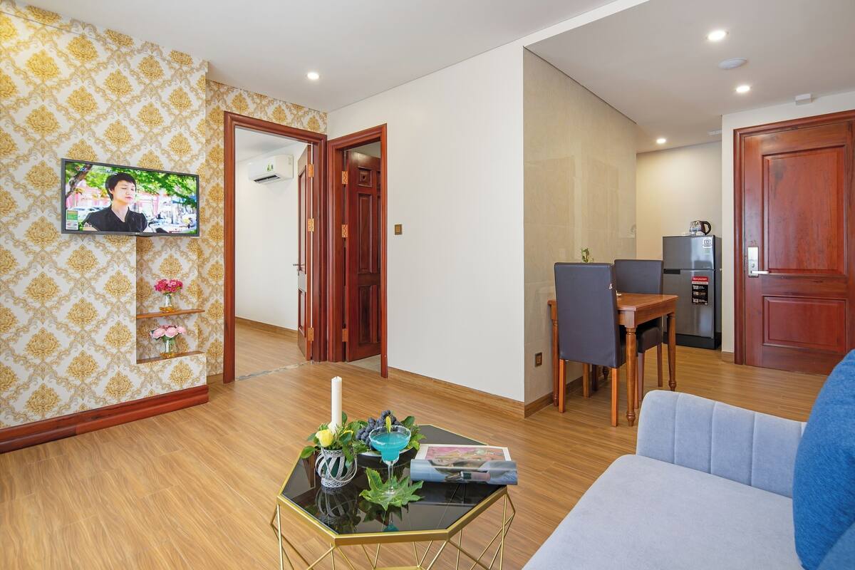 A living room with a TV, a sofa, a coffee table with flowers, and a dining area with chairs and a table.