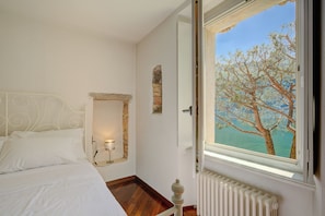 A bedroom with a bed, a nightstand, a wall-mounted lamp, a stone wall feature, a window with a view of trees and water, and a radiator. - Palazzo Gritti Papavero, Morcote, Switzerland (Morcote)