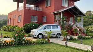 Een tweelaags huis van rode baksteen met een witte auto ervoor, omringd door een tuin met diverse planten en bloemen.