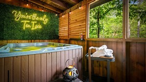 A wooden cabin with a hot tub, a neon sign, a lantern, and a window with a view of trees. - New Cozy Spa Cabin! Hot Tub, Sauna, Jacuzzi! 20% Off-New Listing Promotion! (Sevierville)
