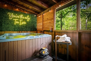 A wooden cabin with a hot tub, a neon sign, a lantern, and a window with a view of trees. - NEW Cozy Couples Spa Cabin! Hot Tub, Sauna, Jacuzzi, 5 Min to Pigeon Forge! (Sevierville)