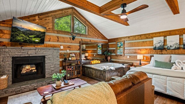 A log cabin interior with a stone fireplace, a hot tub, a large bed, and a loft with a painting. - New Cozy Spa Cabin! Hot Tub, Sauna, Jacuzzi! 20% Off-New Listing Promotion! (Sevierville)