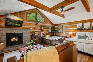 L’intérieur d’une cabane en rondins, avec une cheminée en pierre, un jacuzzi, un grand lit et une mezzanine où est accrochée une peinture.