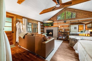A log cabin interior with a fireplace, wooden ceiling beams, a sofa, and a bed. - NEW Cozy Couples Spa Cabin! Hot Tub, Sauna, Jacuzzi, 5 Min to Pigeon Forge! (Sevierville)