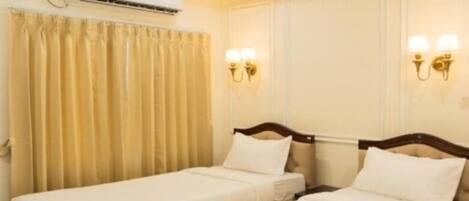 A hotel room with two single beds, a window with curtains, an air conditioning unit, and wall-mounted lighting.