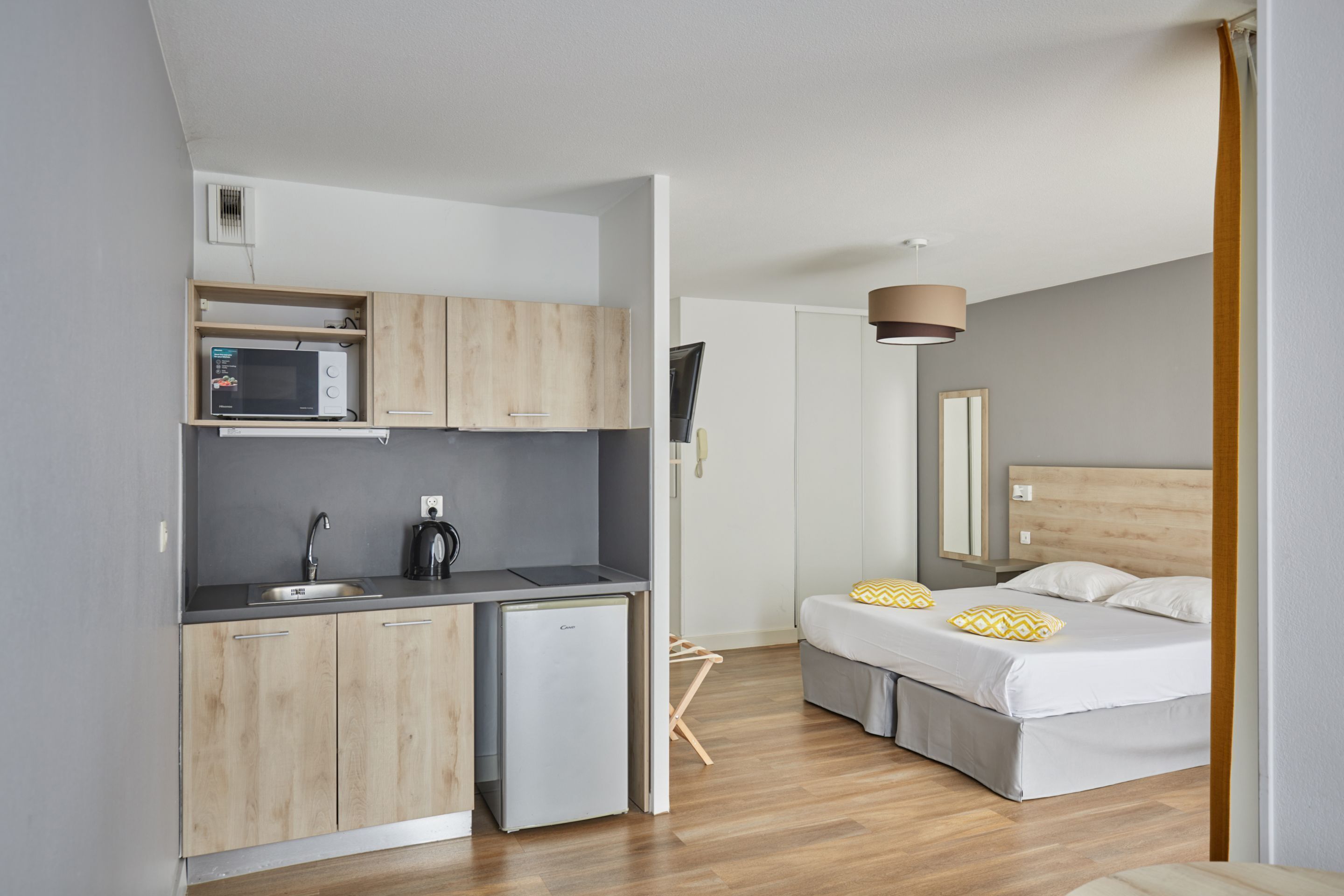 Cocina compacta con microondas, lavamanos y refrigerador, junto a una cama con cabecera de madera y dos almohadas.