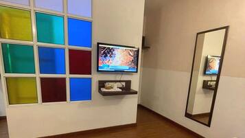 Habitación de hotel con cama, televisor sobre un soporte, espejo y pared con paneles de vidrio de colores.