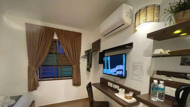 A room with a bed, a desk with a computer, a chair, a window with curtains, an air conditioner, a television, and a shelf with bottles.