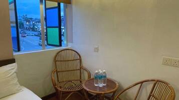 Habitación de hotel con una cama, dos sillones de mimbre, una mesita redonda con botellas de agua, una ventana que da a la calle y una lámpara de pared.