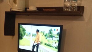 A television mounted on a wall, a kettle, two bottles of water, a roll of toilet paper, and a basket with cards.