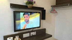 A television mounted on a wall with a shelf below displaying a roll of toilet paper and a decorative item.