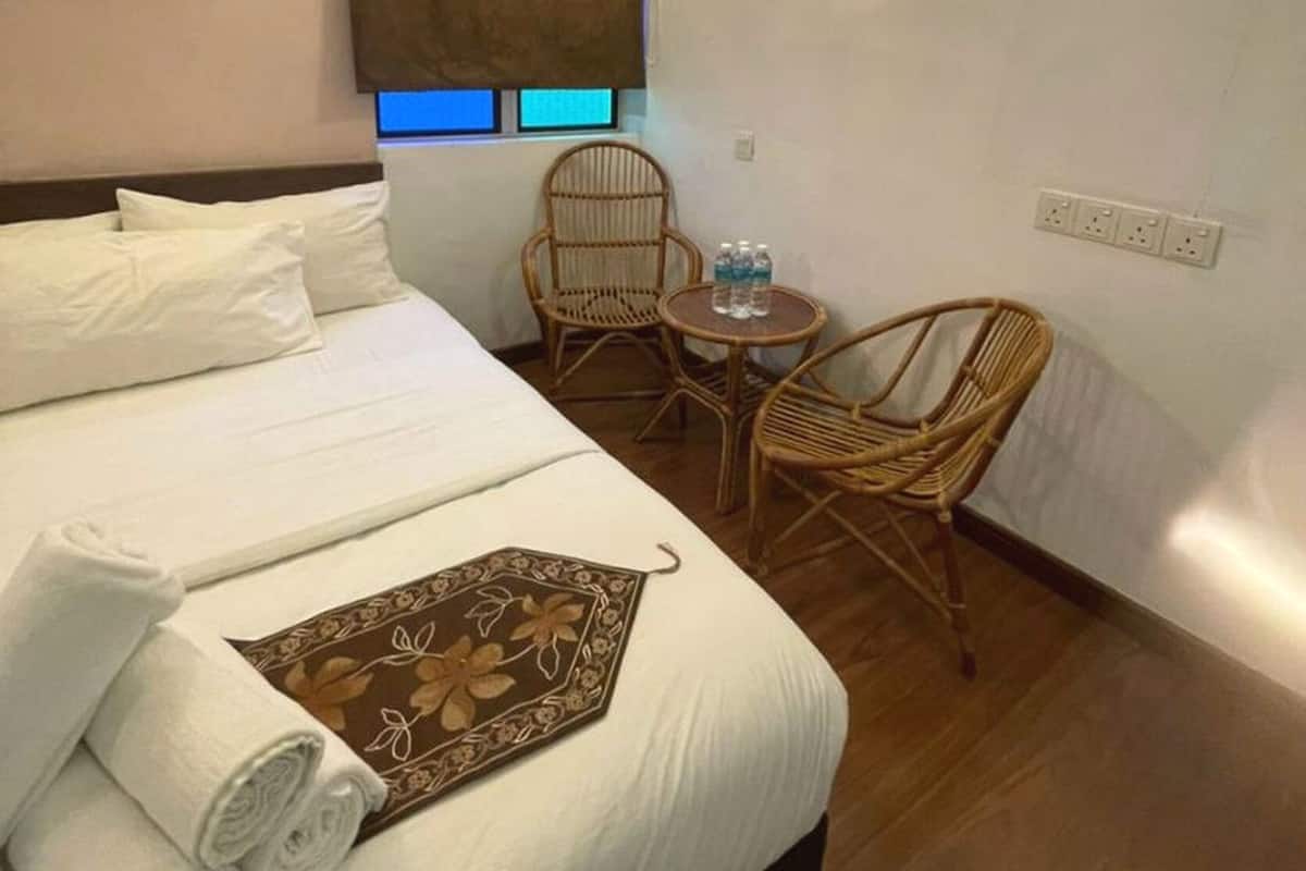 A hotel room with a bed, two wicker chairs, a small table with water bottles, a painting on the wall, and an air conditioning unit.