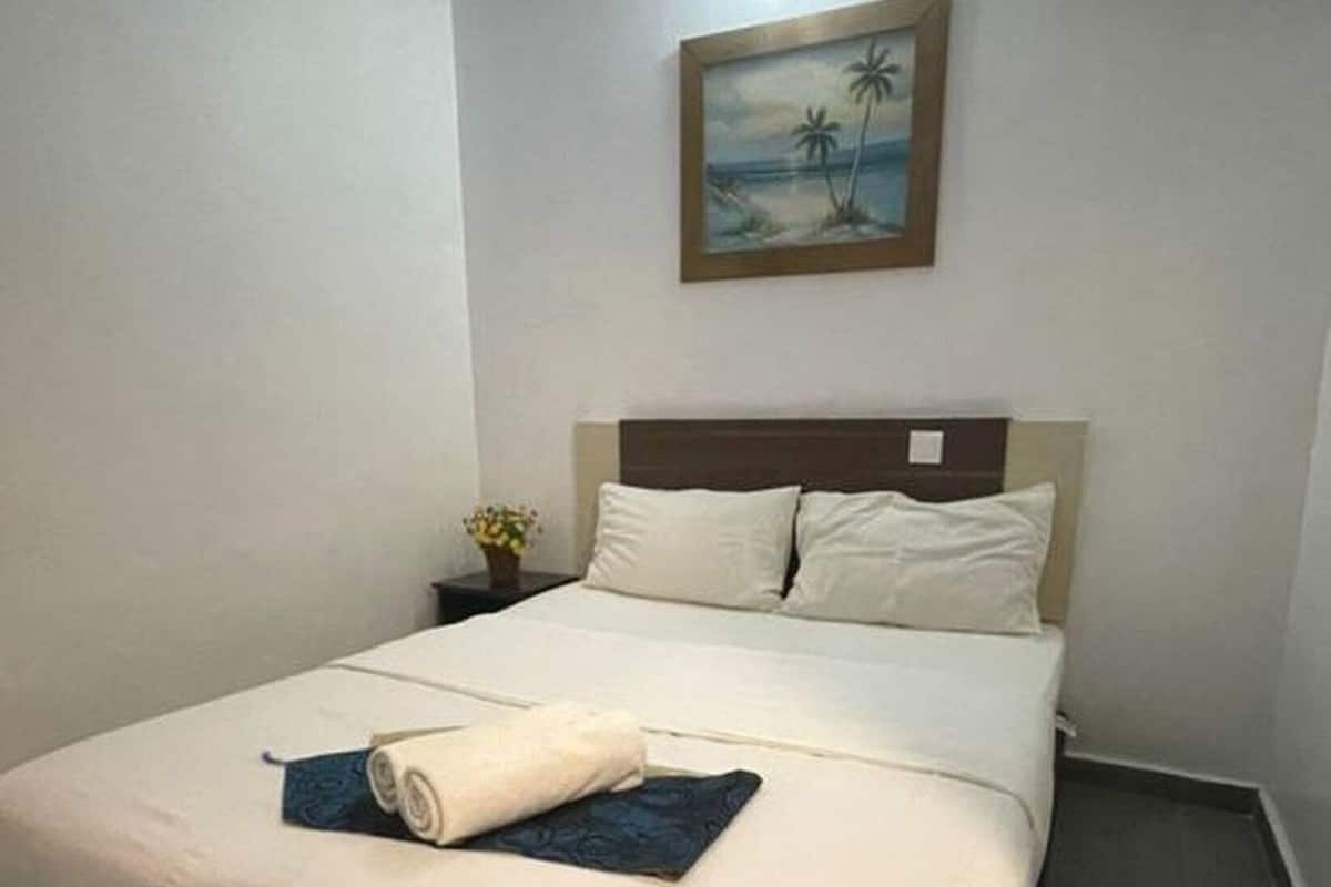 A neatly made bed with a blue towel and a rolled-up towel, a nightstand with a small plant, and a painting of a beach scene on the wall.