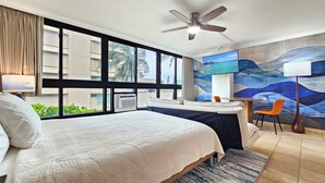 Quarto de hotel com uma cama, uma escrivaninha, uma cadeira, ventoinha no teto e uma janela grande com vista para palmeiras e edifícios.
