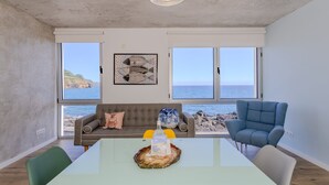 Ein modernes Wohnzimmer mit einem großen Tisch, zwei Sofas, einem blauen Sessel und Meerblick durch große Fenster.