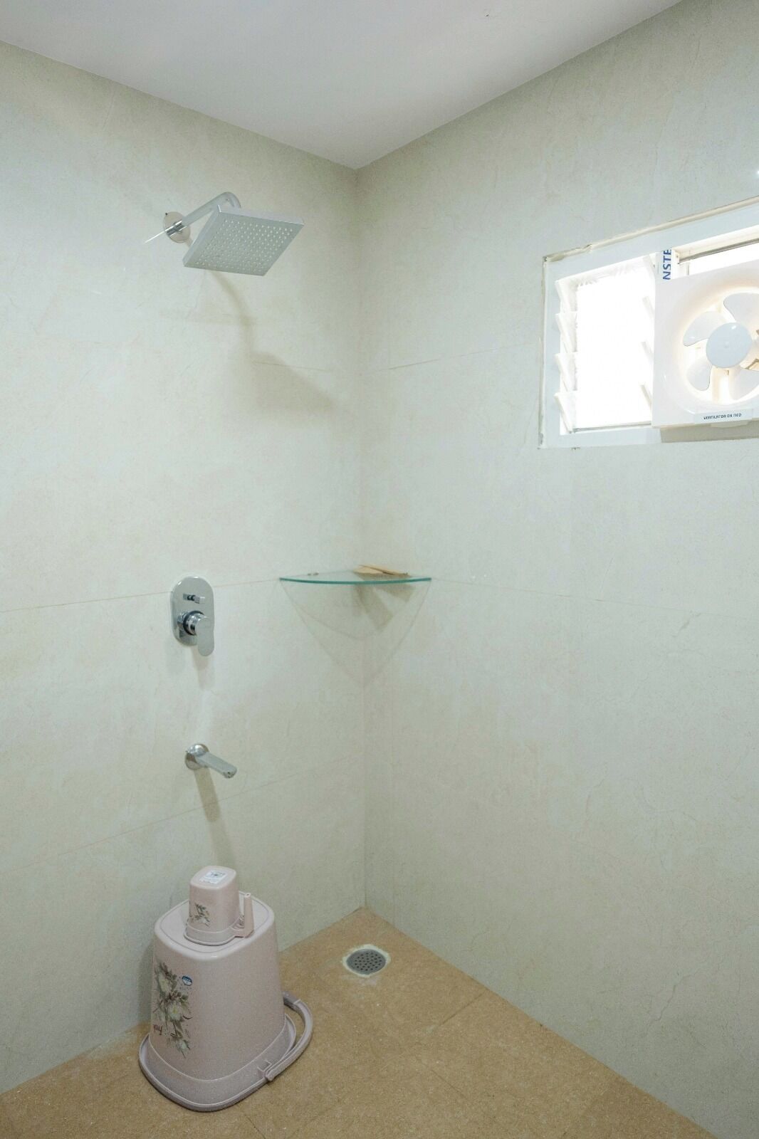 A small, enclosed shower area with a handheld showerhead, a glass shelf, a toilet, and a window with a fan.