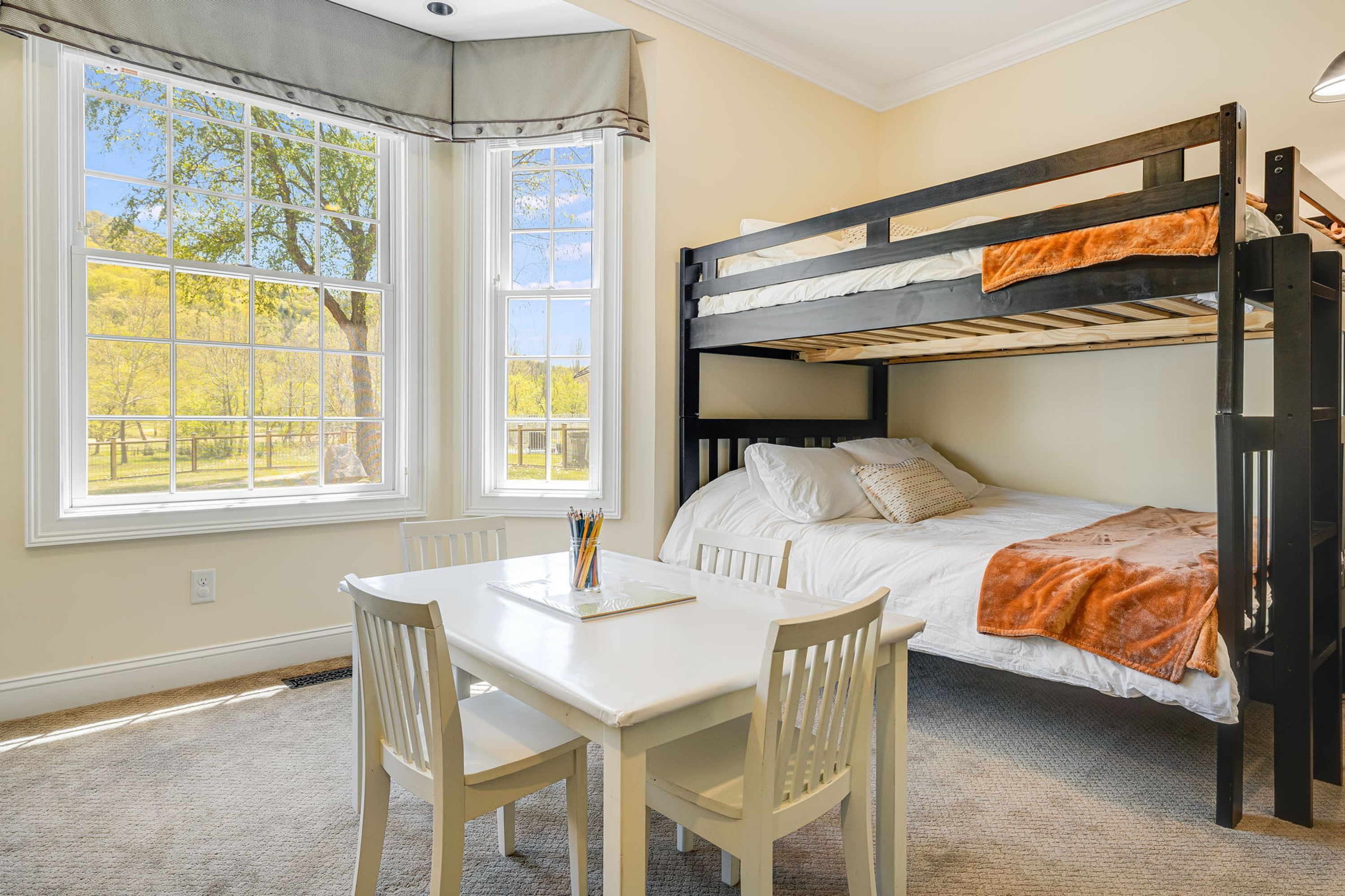 A bunk bed with white bedding, a desk with chairs, and a window with a view of trees.