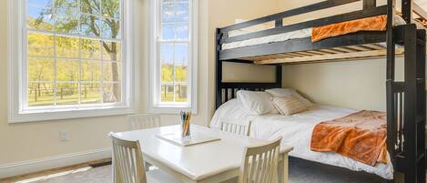 A bunk bed with white bedding, a desk with chairs, and a window with a view of trees.