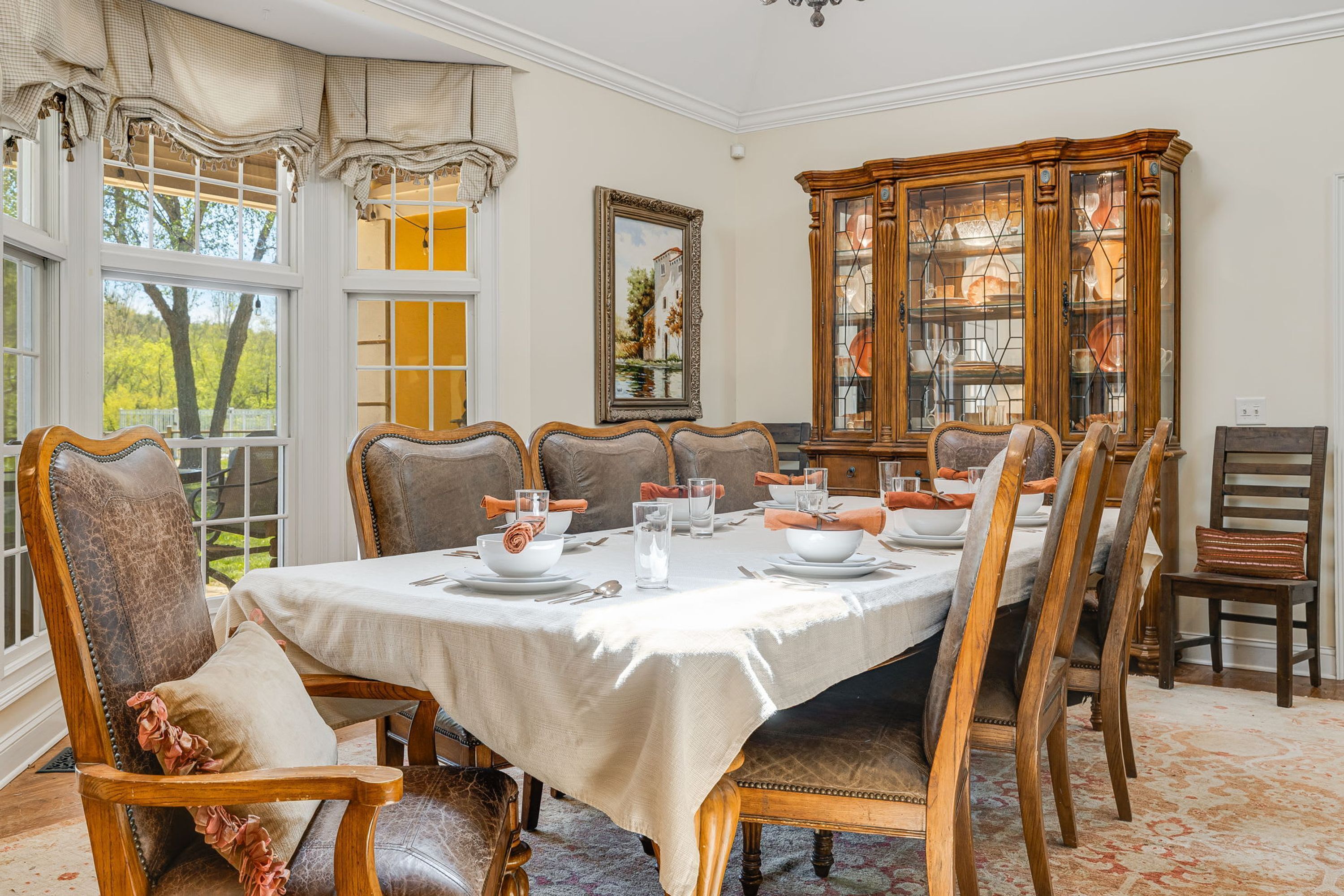 A dining room with a table set for six, surrounded by wooden chairs, a china cabinet, and a large window with curtains.