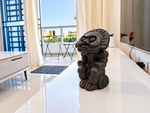 A modern living room with a white sofa, a small table with a decorative sculpture, a flat-screen TV, and a balcony with a view of buildings and greenery.