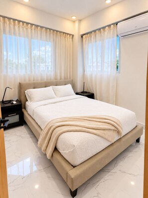 A modern bedroom with a large bed, a nightstand, an air conditioning unit, and sheer curtains. - The Duo by Suarez Properties | Jacuzzi & BBQ (Santo Domingo Este)