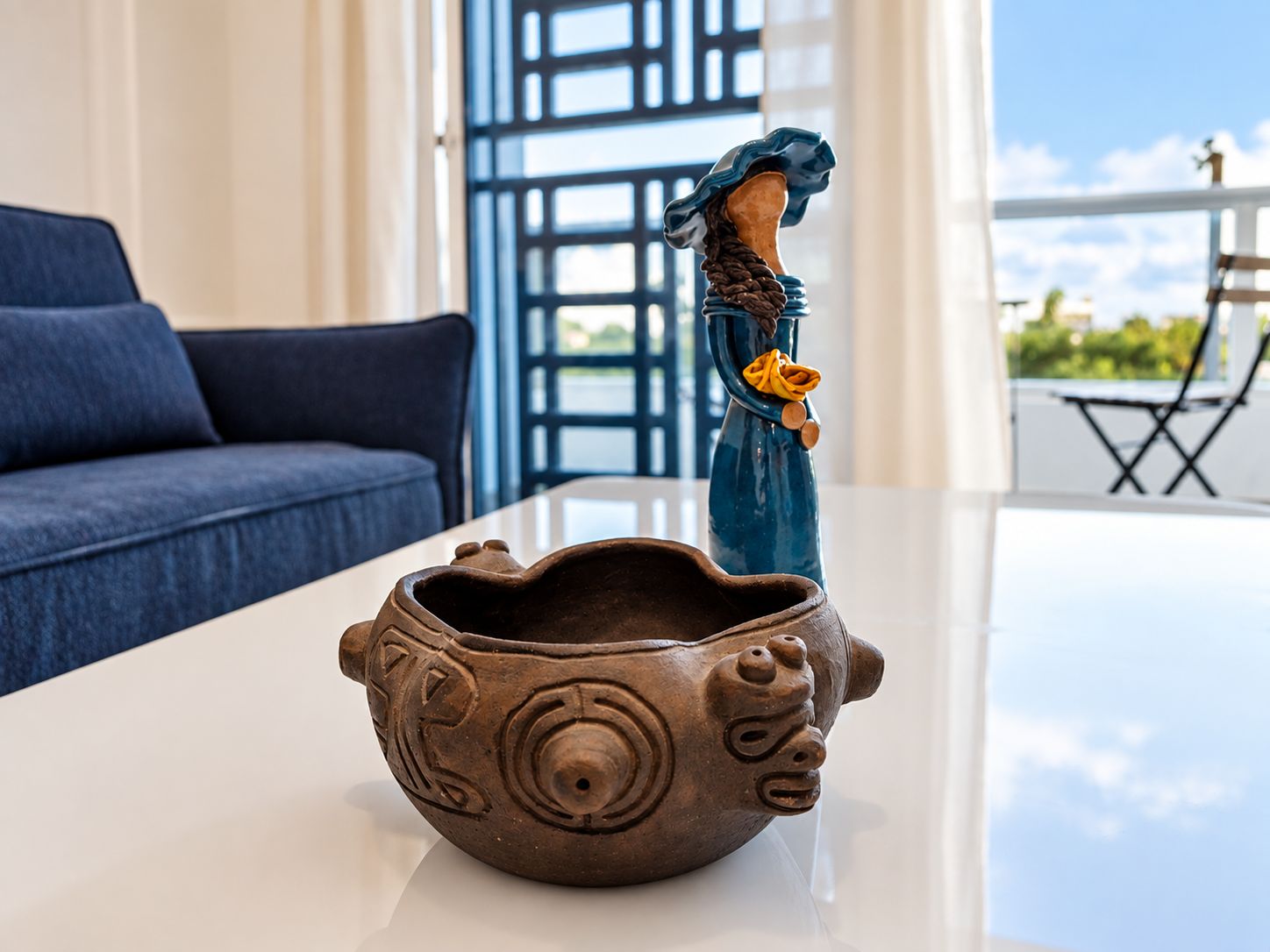 Eine dekorative Keramikschale mit einer Figur auf einem weißen Tisch in einem Zimmer mit einem blauen Sofa und Blick ins Freie.