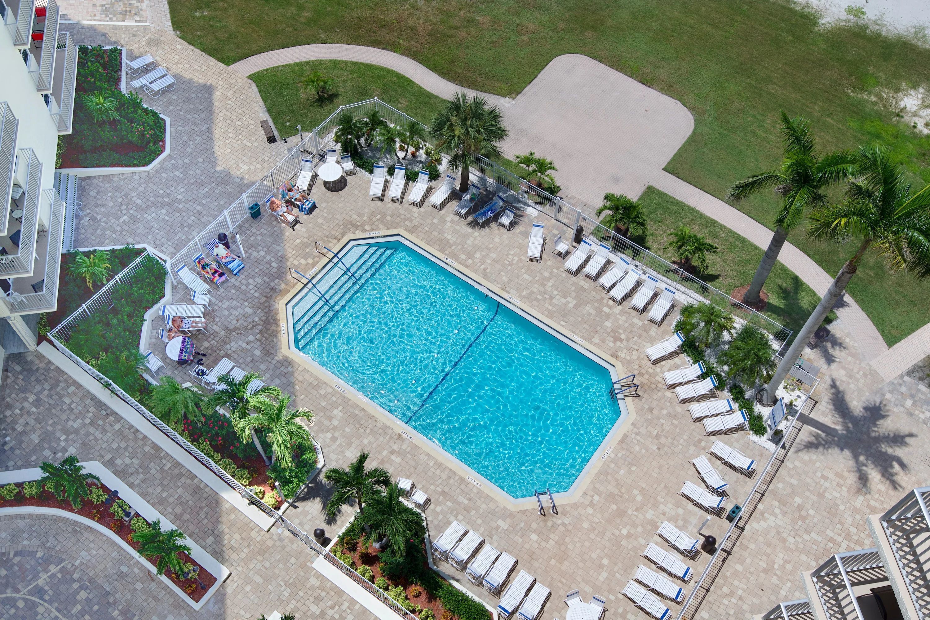 An aerial view of a resort with a central swimming pool, sun loungers, palm trees, and a landscaped garden.
