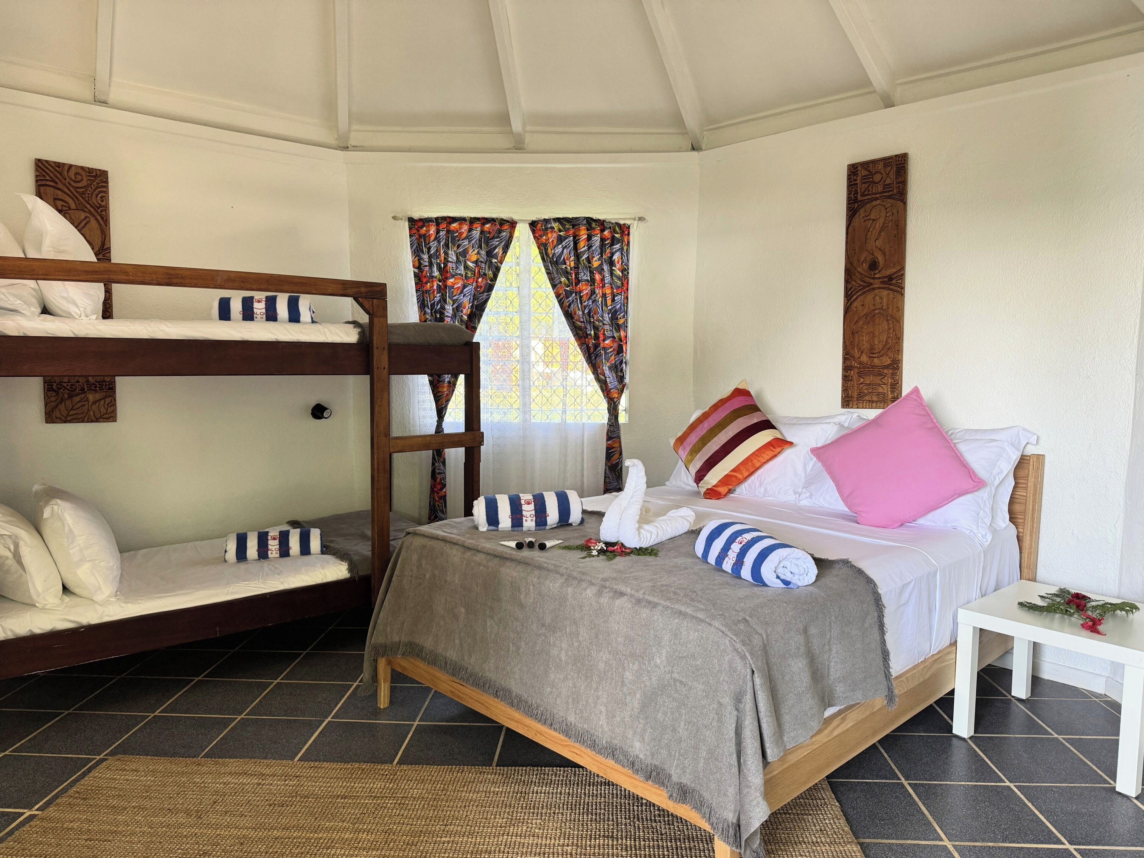 A room with a bunk bed, a single bed with pillows and a blanket, a small table with a flower, and a window with curtains.