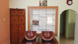 A room with two wooden chairs, a wooden wardrobe, a window with a decorative frame, and a clock on the wall.