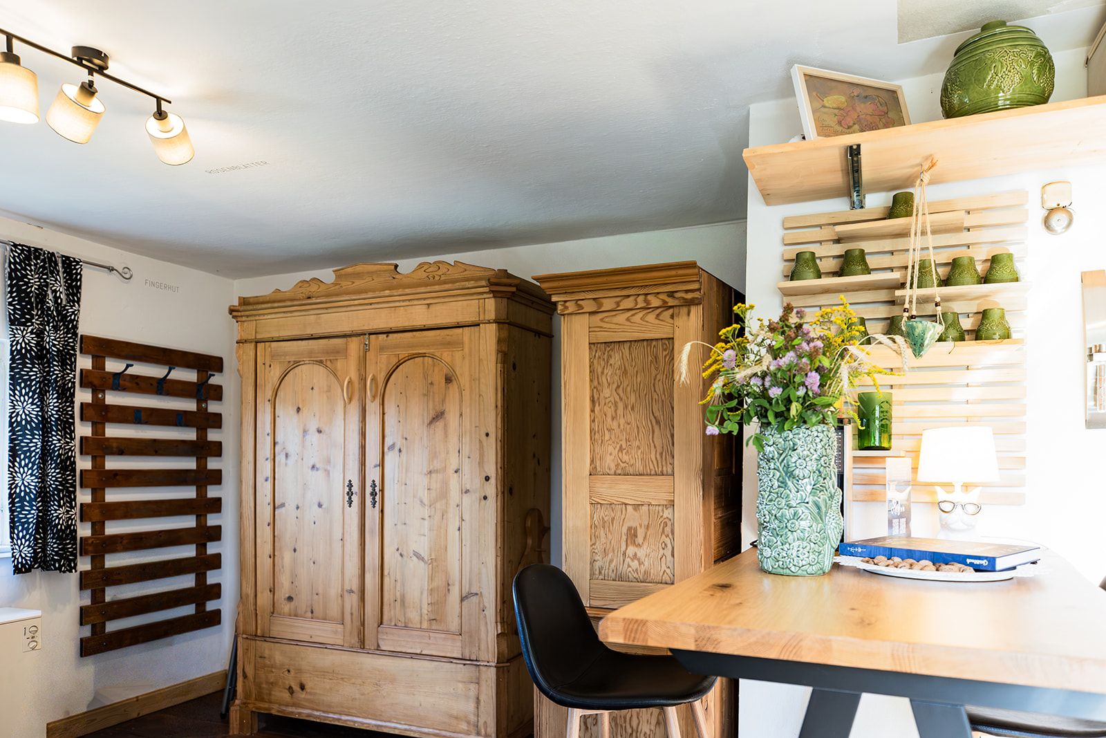 Un armario de madera con tapa de listones, una silla negra, una mesa con un jarrón de flores y una estantería con diversos objetos.