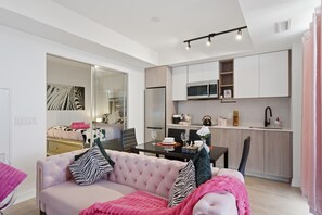 Un salon moderne avec un canapé rose, une table à manger et un coin cuisine doté de meubles blancs et d’un four à micro-ondes.