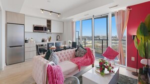 A modern apartment with a kitchen, dining area, and living room. It features a large window with a city view, a pink sofa, and a dining table with chairs.