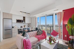 Un appartement moderne comprenant une cuisine, un coin repas et un salon. Il dispose d’une grande fenêtre offrant une vue sur la ville, d’un canapé rose et d’une table à manger avec des chaises.