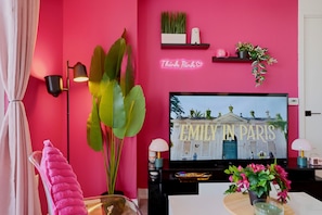 Habitación con televisión mostrando 'EMILY IN PARIS', pared rosa, planta grande y letrero luminoso.