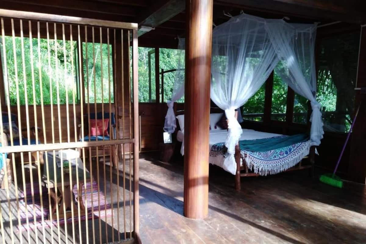 Una habitación de madera con una cama bajo un dosel, una silla de madera y una zona con barrotes.