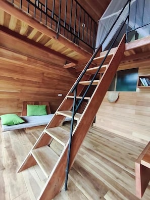 A wooden interior with a staircase, a bed with green pillows, and a small seating area. - Ella Treehouse (Ella)