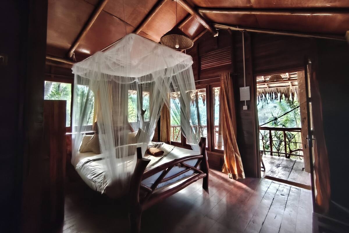 Habitación de madera con cama bajo un dosel, ventanas amplias y balcón con vistas a zonas verdes.
