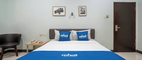 A hotel room with a bed, two pillows with 'rehat' branding, a chair, a small table with a phone, and a door.