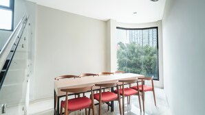 A modern dining area with a long table and chairs, a staircase, and a large window with a view of greenery.