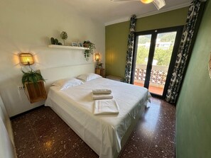 A hotel room with a bed, two bedside tables, a wall-mounted shelf with plants, a window with curtains, and a view of greenery outside. - Condo in Roses · ★4.68 (Roses)